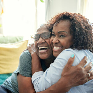 Waist-up view of Black women in early 50s and late 60s sitting on sofa in family home, cheek to cheek and grinning as they embrace.